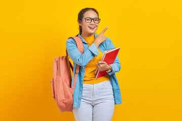 Wall Mural - Portrait of smiling young Asian woman student in casual clothes with backpack holding book and pointing aside with finger isolated on yellow background. Education in college university concept