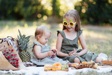 Candid lifestyle portrait of two caucasian siblings eating healthy food on picnic blanket at summer. Cute baby one year old and her sister seven years old having fun together during vacations on