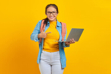 Wall Mural - Portrait of smiling young Asian woman student in casual wear with backpack pointing at laptop with finger isolated on yellow background. Education in college university concept