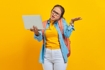 Wall Mural - Portrait of angry young Asian woman student in casual clothes with backpack talking on smartphone while holding laptop,saying no with palms isolated on yellow background