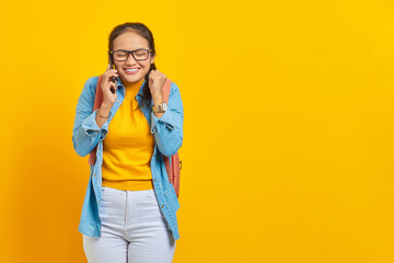 Wall Mural - cheerful young Asian woman student in casual clothes with backpack holding mobile phone, showing celebrating luck gesture isolated on yellow background. Education in college university concept