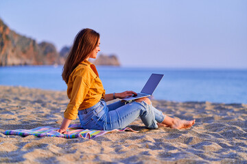 Wall Mural - Young happy joyful smiling carefree satisfied millennial freelancer woman using computer and sitting on sand beach by the sea. Dream office remote work concept