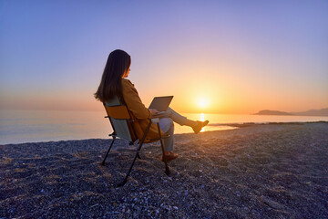 Wall Mural - Satisfied free millennial freelancer woman using computer and sitting on beach by calm sea at sunset. Enjoyment of dream office remote work concept