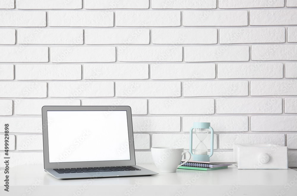 Workplace with laptop, cup and hourglass near white brick wall