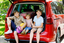 Three Children, Two Boys And Preschool Girl Sitting In Car Trunk Before Leaving For Summer Vacation With Parents. Happy Kids, Siblings, Brothers And Sister With Suitcases And Toys Going On Journey