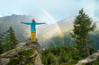 © IBEX.Media - Woman standing with arms outstretched out in front of a beautiful scenery with mountains and rainbow after rain
