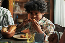 African Little Boy Sitting At Dining Table And Eating Roast Chicken During Dinner With His Family