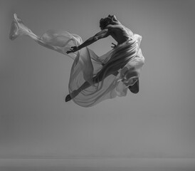 Liberty. Black and white portrait of graceful muscled male ballet dancer dancing with fabric, cloth isolated on grey studio background. Grace, art, beauty concept.