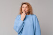 © Red Cristal - Oops. Embarrassed middle age woman with curly blonde hair use hand cover mouth for not commenting, looks with a having mysterious facial expression, stands in trendy t shirt over light grey background