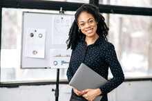 Portrait Of A Positive Pretty Elegant African American Business Woman, Financial Manager Or Mentor, Broker, Marketer, Stand In The Office, Holds Laptop In Hands, Looks At Camera, Smiling Friendly