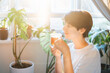 © okrasiuk - Woman with closed eyes drinking tea in a room with green house plants, enjoying its taste and aroma in a sunset or sunrise light. Relaxing and self-care, personal fulfillment. Selective focus.