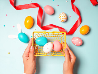 Poster - Top view of hands holding a shopping basket with Easter eggs on a blue background with a red ribbon