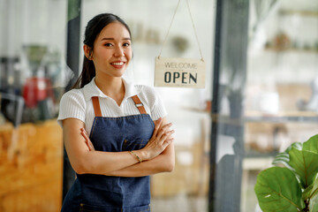 Young Asian businesswoman small business owner standing at cafeteria door entrance pen signboard, A cheerful entrepreneur young waitress in a blue apron near the glass door 