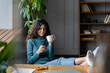 Young relaxed businesswoman drinking coffee and using cellphone at workplace with legs on office table, happy female employee checking social media feed on smartphone, taking break during workday