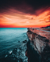 Vertical Shot Of A Man On The Edge Of A Cliff Near The Sea With Turqoise Blue Water And Red Sunset
