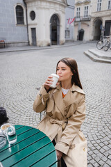 young woman in trench coat drinking coffee to go on cafe terrace.