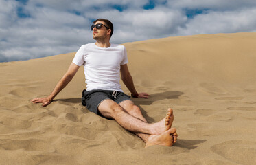 Brunette man wearing white t-shirt sitting on warm sand.