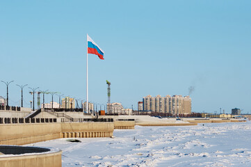 Snow-covered embankment of the Amur River with the state flag of the Russian Federation. Two levels of coastal fortifications. Fences stairs and lanterns.