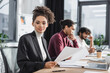 © LIGHTFIELD STUDIOS - Young african american businesswoman looking at camera near laptop and blurred businessmen in office.