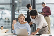 © LIGHTFIELD STUDIOS - Businesswoman holding takeaway drink while using laptop near indian colleague in office.