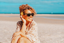 Portrait Of Happy Tourist Sitting And Smiling At The Beach With Blue Sky And Ocean In Background. Travel And Tourism In Summer Holiday Vacation. Female People With Sunglasses On The Sand