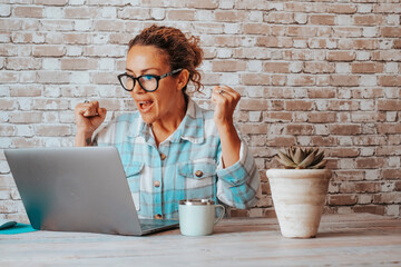 Wall Mural - Happy woman exult and celebrate in front of a laptop after receiving a notification or email message. Overjoyed people working on computer at office or home. Female having an idea about her job