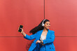 © Dani D.G - Young black woman in her 20s with curly hair, wearing a 90s, 80s retro style blue outfit, dancing hip hop against a red wall, dynamic and fun expression.