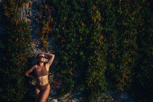 Beautiful Sexy Girl In Sunglasses In A Beige Swimsuit. Young Woman With Tanned Body Soit Near Stone Wall With Green Plants