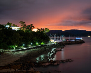 Wall Mural - Lights on the boardwalk to a marina at sunset on a calm ocean. Red sky behind the mountain range.