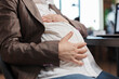 © DC Studio - Close up of pregnant woman touching baby bump while in company office workplace. Future young mother sitting in marketing company workplace while holding baby belly bump.