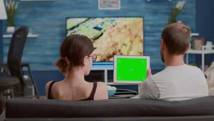 Couple relaxing on sofa looking at social media video content using digital tablet with green screen in living room. Man and woman enjoying free time watching influencer vlog on chroma key device.
