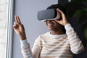 Young smiling african american female in VR glasses shopping in virtual reality, exploring virtual store, picking up and examining items while sitting by window indoors. Augmented Reality in retail