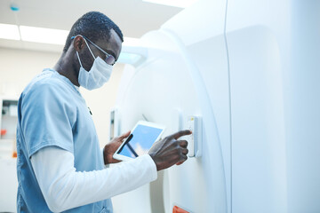 African-American MRI technologist in mask and glasses holding tablet while turning on equipment