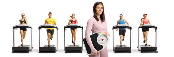 Canvas Print - Young fit woman in sportswear holding a measuring scale and people running on treadmills in the back