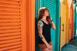 Summertime. Sexy woman with a tattoo on her arm, standing leaning by beach's changing room. Multicolored wooden houses on the background