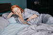 © Beznika - young beautiful red-haired woman dressed in blue pajamas lies on the bed and uses a smartphone