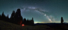 Panoramic View Of Evening Mountain Hills Under Beautiful Starry Sky. Small Flame, Silhouette Of Tent Near Trees On The Background Of Fabulous Landscape Of Milky Way Galaxy And Snow-capped Mountains.