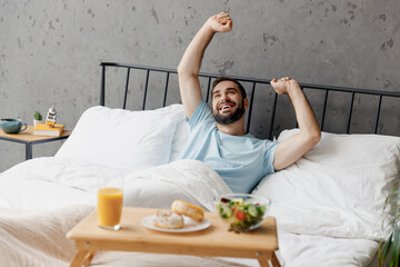 Wall Mural - Young happy fun man wear blue t-shirt lying in bed stretch hands look at breakfast table hands rest relax spend time in bedroom lounge home in own room house wake up dream. Good mood bedtime concept.