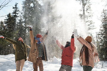 Group of young playful men and women enjoying spending time outdoors on winter day throwing snow in the air