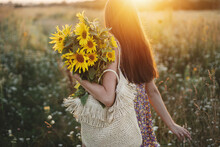 Beautiful Woman Holding Sunflowers In Warm Sunset Light In Meadow. Tranquil Atmospheric Moment In Countryside. Stylish Young Female In Floral Dress Walking With Sunflowers In Summer Evening Field