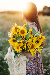 Wall Mural - Beautiful woman holding sunflowers in summer evening meadow. Tranquil atmospheric moment in countryside. Stylish young female in floral dress walking with sunflowers in warm sunset light in field
