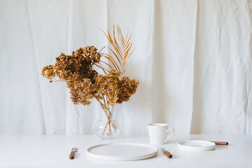 Photo of a minimalistic white simple dinnerware cup plate and glass flower vase in modern style on sunlight indoors