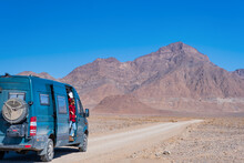 Woman In A Red Checkered Sweater Peeking Out Of A Campervan With A Dusty Road Ahead