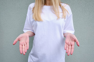 Wall Mural - Blond hair woman posing near a light wall. Beautiful young caucasian girl. Hand gestures. Emotion. Casual clothing. Studio model in work. Strong woman, future is female. Jeans and white blank t-shirt
