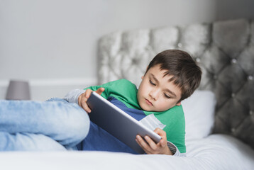 Wall Mural - Young boy using tablet playing game on internet with friends, Homeschooling Kid doing homework online by digital pad at home, Child lying on bed relaxing,watching cartoon or talking online with friend