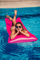 Wall Mural - Sexy woman in a swimsuit lies on a pink inflatable mattress in the pool. Relax by the pool on a hot summer sunny day. Vacation concept