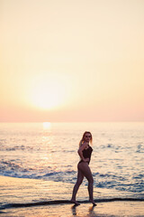 Wall Mural - Beautiful lady in a black swimsuit on the seashore, Smiling face, with a straw hat on her shoulders. Golden sunset light