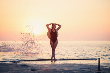 Wall Mural - Attractive young girl with long hair poses in front of the camera on the beach. She is wearing a black swimsuit. Golden sunset light