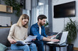 © Liubomir - Young family, husband and wife, reviewing their bills, loan and mortgage agreements, sitting on the couch at home, working with documents on a laptop, embarrassed, upset