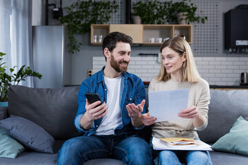 A man and a woman are happy at home, make online payments, using the phone and banking application, sitting on the couch and holding bills